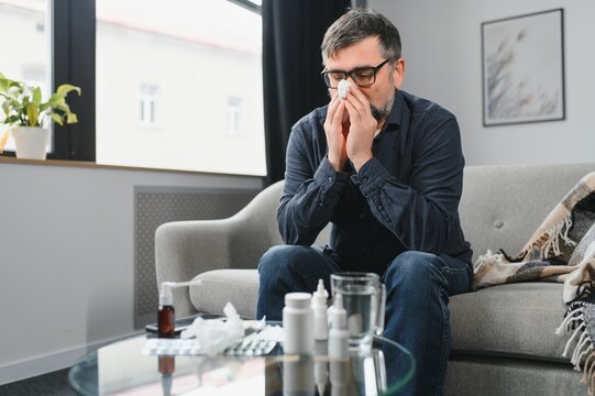 Healthcare, Flu, Hygiene And People Concept - Sick Senior Man With Paper Wipe Blowing His Nose At Home