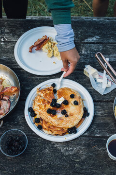 Camping Breakfast: Woman Serving Stack Of Sliced Pancakes, Scrambled Eggs, Bacon On Picnic Table