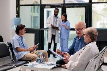 Obraz premium Senior specialist sitting on chair in hospital lobby explaining illness diagnosis to elderly patient discussing health care treatment. Diverse people waiting to start examination during checkup visit