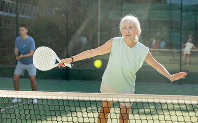 Sporty mature woman padel player hitting ball with a racket on a hard court