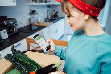 Online home food delivery. Focus on active app with order list on the phone screen. Woman checking her order. Cardboard box with fresh vegetables standing on the kitchen table. Local farmer food.