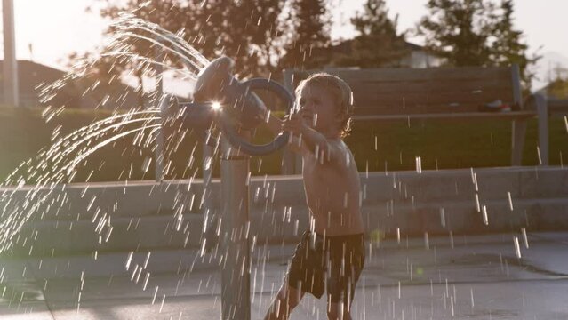 3 year old boy joyfully spins around with large splash pad water feature, sunset