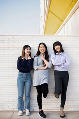 Vertical photo of a happy group of young Asian women playing with each other outside. The 3 girls are leaning against a wall as they look at the camera smiling.Urban lifestyle concept