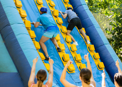 Two male friends having fun in outdoor amusement park on summer day, climbing on inflatable slide with wooden poles..