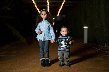 Two dark-skinned children posing outdoors at night holding hands while looking at the camera, the...