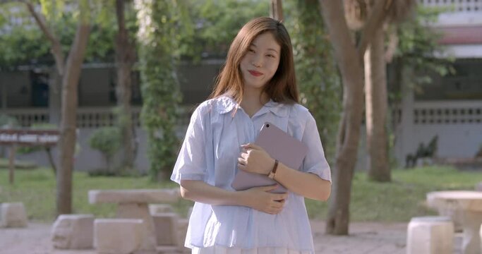 (Slow motion)A happy smiling Asian woman who is a Chinese teacher and teaches Chinese language in a secondary school of Thailand, standing with her tablet in the school park in the evening.