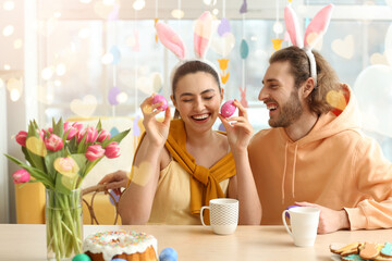 Happy couple in bunny ears and with Easter eggs sitting at table in kitchen
