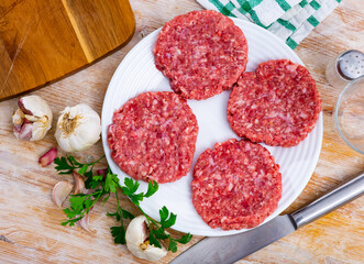 Shaped patties for burgers from raw ground beef meat with fresh parsley sprigs, spicy garlic and allspice on plate. Cooking ingredients