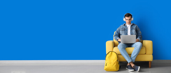 Male student with laptop studying in armchair near blue wall. Banner for design