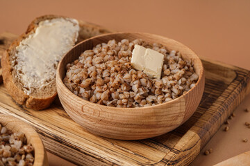 Wooden bowl of tasty buckwheat porridge with butter and bread on beige background
