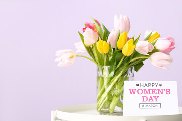 Vase with tulips and greeting card for Women's Day on table near lilac wall