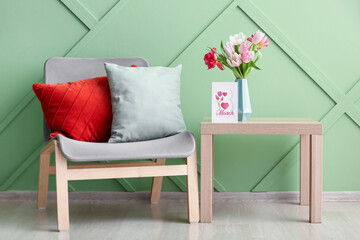 Vase with tulips, greeting card for Women's Day on table and chair near green wall in room