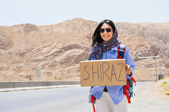 Young traveler woman stand on side of road hitchhike with sign to Shiraz and thumbs up with smile in Iran .Solo safe travel in Asia concept