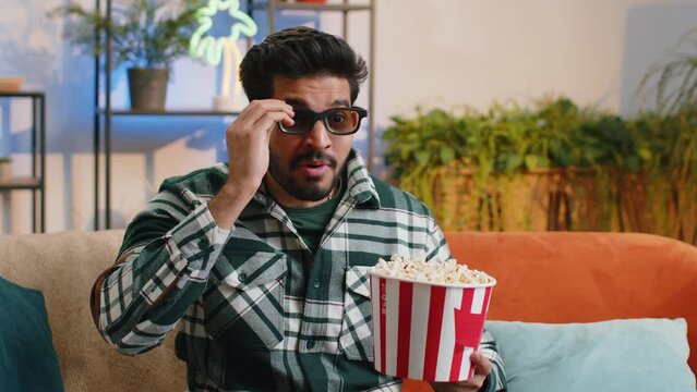 Excited indian man in 3D glasses sits on couch eating popcorn snacks and watching interesting TV serial sport game, film online social media movie content at home apartment. Guy enjoying entertainment