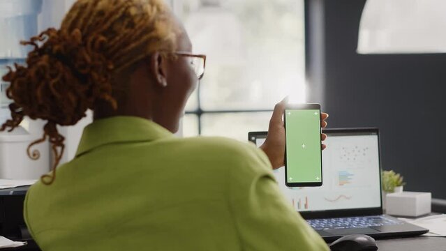 Female manager looking at greenscreen display in office, working with isolated mockup template on mobile phone. Young employee using blank copyspace mockup in coworking space.
