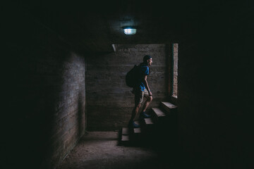 Young latin man with a bag coming out of the stairs of a basement going to a place with natural light. - obrazy, fototapety, plakaty