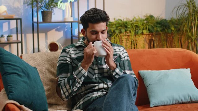 Handsome smiling indian man drinking a cup of warm coffee or herbal tea at home living room apartment. Break time. Hindu guy enjoying comfortable relaxing sitting on couch in the morning. Lifestyles