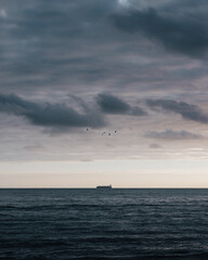 In the vast expanse of the ocean, a lone cargo ship cuts through the waves, its silhouette a stark contrast against the moody twilight sky.