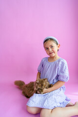 Pretty elementary girl sitting down while holding fluffy brown cat on her lap in front of purple backdrop