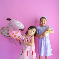 Two cute girls standing while holding a pink animal ballon and baby doll