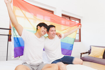 Happy Asian LGBT couple, sitting on the sofa holding and waving rainbow LGBT Pride flag together in the bedroom at home. Diversity of LGBT relationships. A gay couple concept. Loving gay couple.