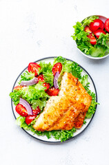 Fried cod fish with salad garnish from lettuce, cherry tomatoes and red onion with sesame seeds, white table background, top view