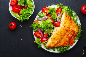 Fried cod fish with salad garnish from lettuce, cherry tomatoes and red onion with sesame seeds, black table background, top view