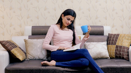 Beautiful Indian girl spending leisure time in reading her favorite novel at home. Medium shot of an attractive young female reading a book while having a cup of tea or coffee - lifestyle concept