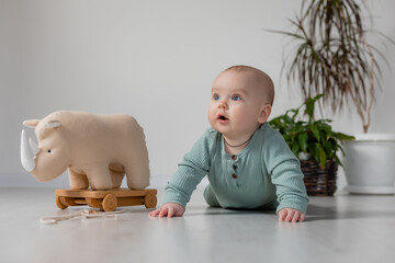 chubby baby in a green jumpsuit is lying on the floor and playing with a wooden toy on a string