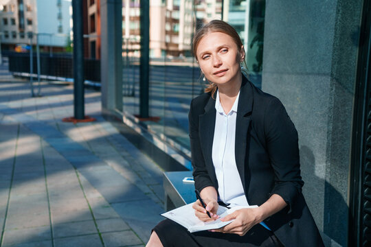 Business woman in suit is in city center against backdrop building, writes, writes down, takes notes, keeps a pen and notebook, brainstorms, creates ideas.