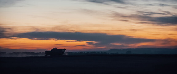 Silhouette of harvester machine to harvest wheat on sunset. Combine harvester driving on field on sunrise. Beautiful dawn sky above wheat field. Combine working in dusk. Wonderful twilight landscape.