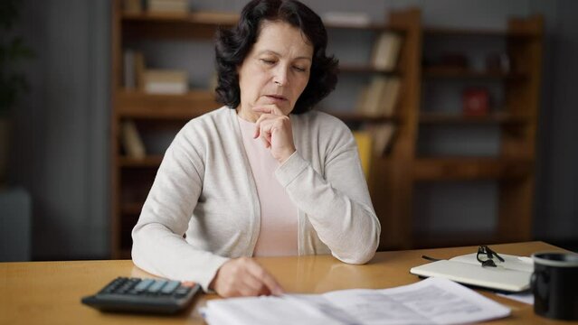 Elderly woman sitting at a table, looking distressed. She is upset about having to make payments on her credit and bills. Reality of many seniors who face financial difficulties. Lifestyle concept.