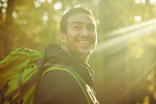 Man, backpacker or hiker in sunshine forest, trekking woods or nature trees in adventure, morning exercise or Japanese fitness. Smile, happy or hiking in sunrise environment, lens flare or wellness