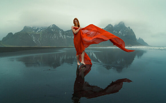 Barefoot lady with red fabric on Reynisfjara beach scenic photography. Picture of person with mountains on background. High quality wallpaper. Photo concept for ads, travel blog, magazine, article