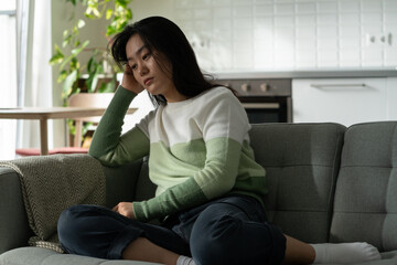 Anxious Asian millennial girl sitting on sofa feeling lonely, suffering from monophobia. Sad depressed young Korean woman sits alone at home looking aside, having problems dealing with panic attack