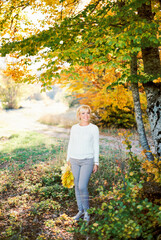 Smiling woman with a bouquet of yellow leaves stands by a tree in an autumn park