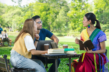 university students learning and doing research together on weekend in the park,group of friends sitting and discussing work, there are computer laptops, text books,coffee cups on table