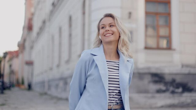 Happy cool blonde woman wearing headphones dancing alone on street. Smiling young beautiful lady listening music standing in city outdoors, feeling free and funky.