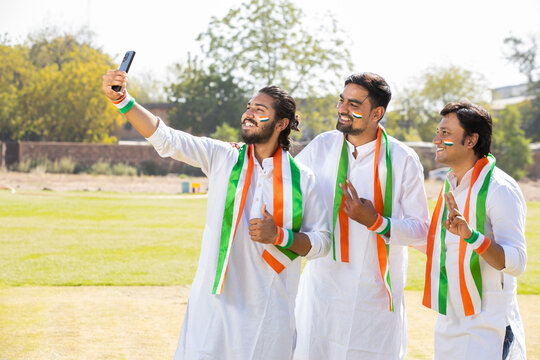 Group Portrait of young indian men wearing traditional white kurta and tricolor duppata taking selife picture with smart phone at park. celebrating Independence day or Republic day.