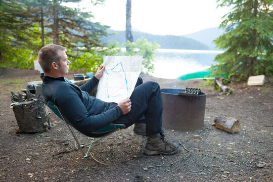 A man reads a map while sitting by a campfire.