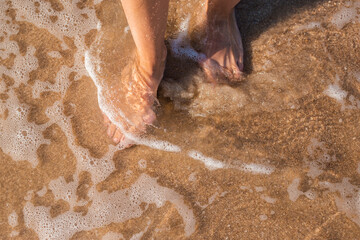 Fototapeta premium Female bare feet stand in the sea on a sandy beach. Top view, flat lay