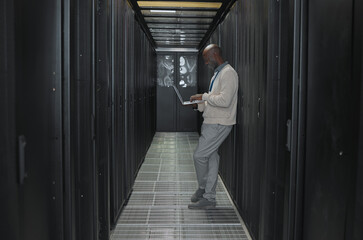 Laptop, network and data center with IT black man for research, engineer working in dark server...
