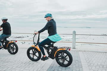 Fototapeta premium Couple, helmet or electrical bike by beach, ocean or sea in future transport, clean energy or sustainability travel. Technology, electric or eco friendly bicycle for happy woman or mature cycling man