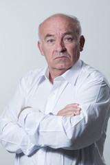 Confident senior man in white shirt crossing hands on chest and looking at camera while standing against gray background. Self confident senior isolated white studio shoot.