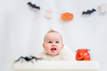 The baby toddler is sitting behind a high chair surrounded by Halloween attributes: pumpkins,...