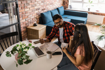 Serious wife and husband planning budget, checking finances, focused young woman using online calculator and counting bills or taxes, man using laptop, online banking services. Family sitting at table