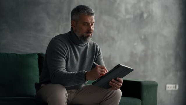 Senior Handsome Man Sitting On Green Couch In Loft Living Room, Working From Home On Tablet Computer