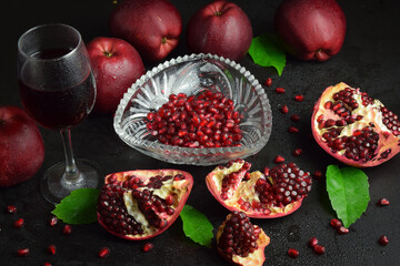 Composition glass of red wine or juice, ripe pomegranates, apples, green leaves indoors. Fresh pomegranate arils. Split open fruit scattered grains seed on black background. Studio shot. Still life