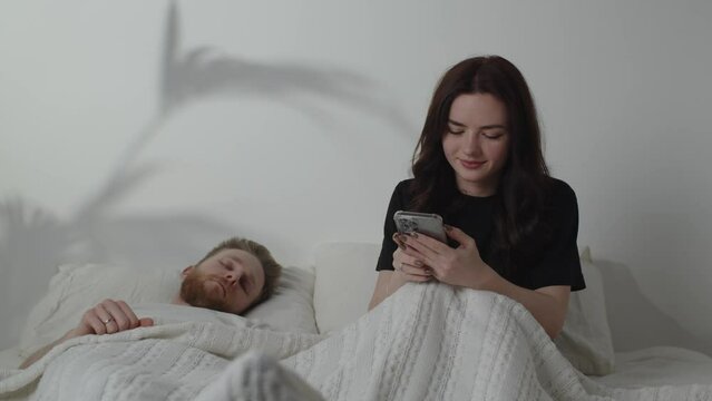 Young woman texting while her husband is sleeping. Beautiful girl typing message on smartphone and looking back at her boyfriend. Cheating concept
