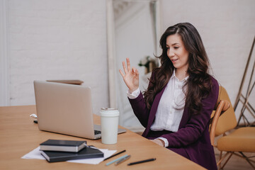 Fototapeta na wymiar Satisfied brunette hispanic woman in violet suit sitting at desk with laptop and cup of coffee makes video call shows ok sign gesture. Cheerful successful entrepreneur celebrates win. Success